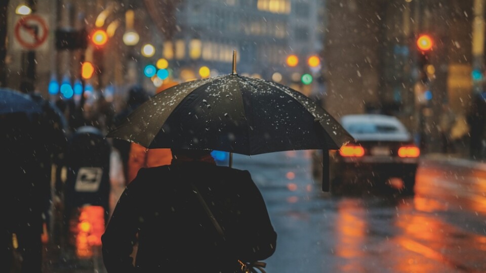 Man in the foreground with umbrella. In the background is a dark and partially lit cityscape.